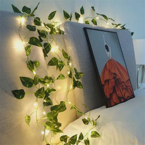 Puede incluir: Una guirnalda de hojas de hiedra artificiales verdes entrelazadas con pequeñas luces LED de color blanco cálido. La enredadera está colocada sobre un cabecero tapizado en gris claro junto a un cuadro de una figura con un vestido naranja.
