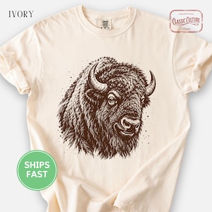 May include: An ivory t-shirt featuring a brown graphic illustration of a bison head in the center. The shirt includes the text IVORY in the top left corner and a logo in the top right corner that reads CLASSIC CULTURE STUDIO.