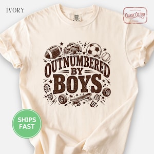 May include: An ivory short-sleeve t-shirt featuring a brown graphic print with the text "OUTNUMBERED BY BOYS". The design includes illustrations of a football, a monster truck, a football, a baseball, a skateboard, and boot prints.