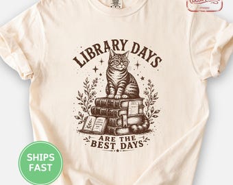 May include: An ivory t-shirt featuring a brown graphic of a cat wearing glasses sitting on a stack of books. The text reads "LIBRARY DAYS ARE THE BEST DAYS" with floral accents and stars surrounding the central illustration.