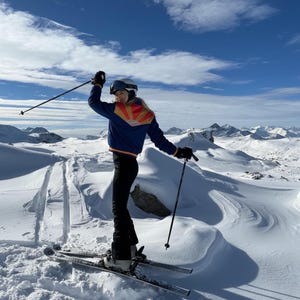 Könnte beinhalten: Eine marineblaue Skijacke mit einer rot-gelben Sonnenaufgangsgrafik auf dem Rücken, schwarze Skihose, ein weißer Helm mit Skibrille und schwarze Skistöcke. Die Kleidung wird auf Skiern in einer verschneiten Berglandschaft unter blauem Himmel getragen.