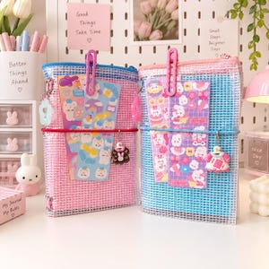Könnte beinhalten: Zwei rechteckige, transparente Netz-Reißverschlusstaschen mit Gittermuster. Die linke Tasche ist rosa mit blauem Rand und rotem Gummiband, die rechte Tasche ist blau mit rosa Rand und blauem Gummiband. Beide enthalten Aufkleberbögen und kleine Anhänger.