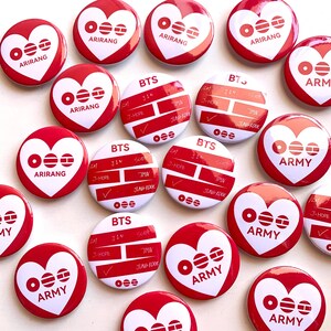 May include: A collection of round pinback badges featuring red and white designs. Some badges display a white heart with red geometric symbols and the text ARIRANG or ARMY. Others show the text BTS with names like JIN, J-HOPE, and JUNGKOOK.