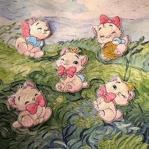 May include: Five embroidered patches featuring white kittens with pink bows. One kitten wears a blue shirt, one holds a yellow ball of yarn, one wears a gold crown and a blue cape with a jewel, one has a pink bow, and one wears a gold crown.