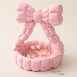 Puede incluir: Un joyero de color rosa claro con textura acolchada y un asa grande en forma de lazo. La base circular contiene un anillo dorado con una piedra transparente, un par de pendientes de perlas blancas y un collar de cadena dorada fina.