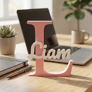 Könnte beinhalten: Ein freistehender dekorativer Buchstabe L in mattem Hellrosa, auf dessen Vorderseite der Name Liam in weißer Schreibschrift angebracht ist. Das Objekt verfügt über einen flachen Sockel, der für einen stabilen Stand auf Oberflächen sorgt.