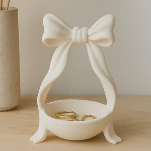 Pode incluir: Um porta-joias de cor creme com uma base em forma de taça, sustentada por duas pernas curvas que se estendem para cima para formar um grande laço decorativo no topo. O objeto de acabamento liso e mate contém vários anéis de metal dourado.