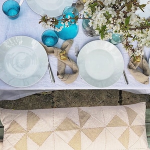 May include: A table setting with white plates, blue glasses, and linen napkins. Each place setting has a small gold bunny rabbit figurine as a napkin ring. The table is covered in a white tablecloth and there are white flowers in a vase in the background.