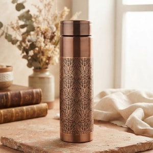 Könnte beinhalten: Eine zylindrische Wasserflasche aus Kupfer mit polierter Metalloberfläche an den oberen und unteren Abschnitten, einem komplizierten, sich wiederholenden floralen Damastmuster in der Mitte und einem passenden flachen Kupferdeckel.