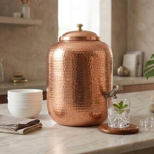 May include: A large, cylindrical, hammered copper water dispenser with a matching lid and a metal spigot. The dispenser features a textured, dimpled surface and a small, light-coloured knob on top of the lid, positioned on a marble countertop.