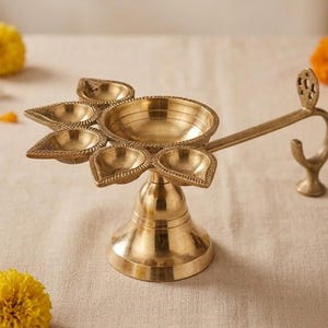 May include: A brass oil lamp featuring a central circular bowl surrounded by five smaller petal-shaped reservoirs. The lamp stands on a flared pedestal base and includes a long, curved handle with a decorative cutout at the top end.