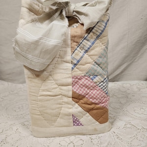 May include: A rectangular fabric pouch featuring a quilted side panel with diagonal strips of pink, blue, and tan plaid and gingham fabric. The main body is cream-colored with stitched leaf patterns and a tied beige and white striped fabric bow closure.