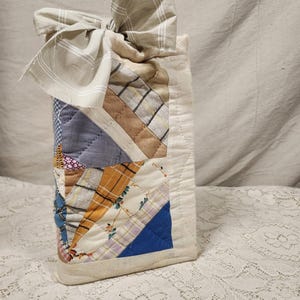 May include: A rectangular fabric pouch featuring a patchwork design of various cotton textiles in shades of blue, tan, yellow, and white. The pouch is topped with a large, tied fabric bow made from a beige and white plaid material.