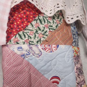 May include: A patchwork quilt featuring various fabric patterns including red floral, green botanical, striped, and light blue prints. The quilt is topped with a white eyelet lace trim and a light pink and white gingham fabric border with raw edges.