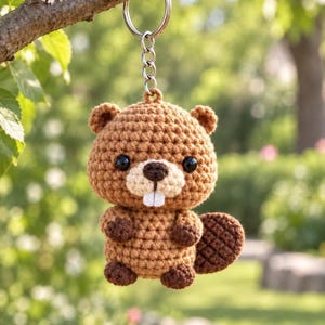 May include: A crochet beaver keychain featuring a light brown body, dark brown paws, and a textured dark brown tail. The beaver has black plastic eyes, a dark brown nose, and two white felt teeth. It is attached to a silver metal split ring.
