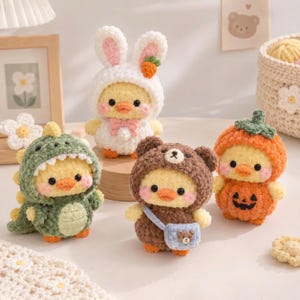 Pode incluir: Quatro pequenas pelúcias de pato amarelas de crochê vestindo diferentes fantasias: um capuz de coelho branco com uma cenoura, um traje de dinossauro verde com espinhos, um capuz de urso marrom com uma pequena bolsa e um traje de abóbora laranja.