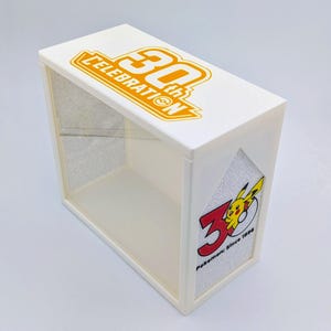 May include: A white rectangular display case with a clear front panel. The top features a yellow 30th Celebration logo. The side panel displays a graphic with the number 30, a yellow Pikachu character, and the text Pokemon: Since 1996.