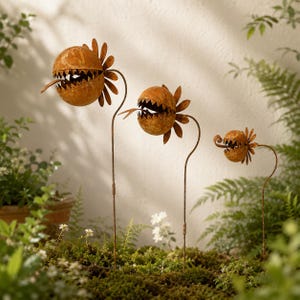 May include: Three rust-coloured metal garden stakes shaped like carnivorous plants with spherical heads, jagged teeth, and protruding tongues. Each plant is mounted on a thin, curved metal stem, arranged in a descending size order from left to right.