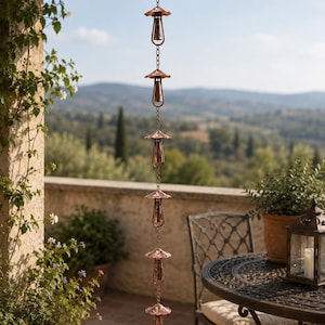 May include: A copper rain chain featuring a series of six vertically linked bell-shaped cups, each topped with a fluted, mushroom-like canopy. The metal chain is suspended from a roof structure, showcasing a metallic finish and a repeating design.