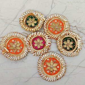 以下が含まれることがあります： 中央に金色の金属製の花が施され、その周りを銀色の尖った金属製の花びらが囲んでいる6つの円形の布製パッチ。中心部はオレンジ、緑、ピンクの色合いで、全体に小さな金属製のアクセントが散りばめられています。