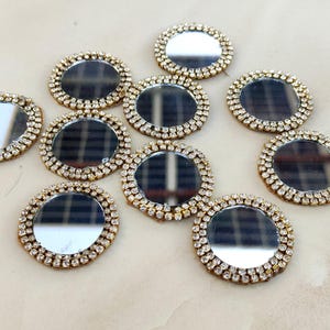 May include: A collection of ten circular mirrors, each featuring a reflective center surrounded by a border of small, clear, faceted rhinestones set in a gold-toned metal frame. The mirrors are arranged in a scattered pattern on a light surface.