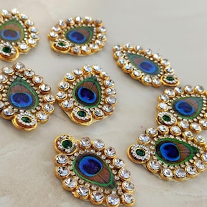 May include: A collection of seven teardrop-shaped appliqués featuring central peacock feather patterns in blue, green, and brown. Each piece is bordered by clear, faceted rhinestones set in gold-toned metal with a small green accent stone.