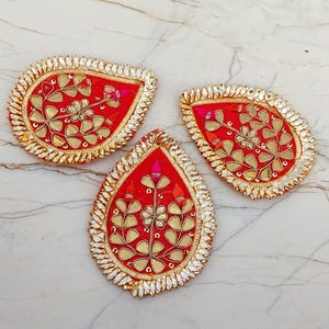 May include: Three teardrop-shaped fabric patches in vibrant red, each featuring gold-toned metallic leaf embroidery and a central floral motif. The edges are bordered with reflective, silver-colored, pointed mirror-like embellishments.