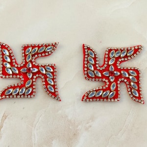 May include: Two red fabric swastika-shaped appliqués decorated with clear marquise-cut rhinestones along the edges and centre. Each piece features a single red circular rhinestone at the centre point, surrounded by clear stones in a geometric pattern.