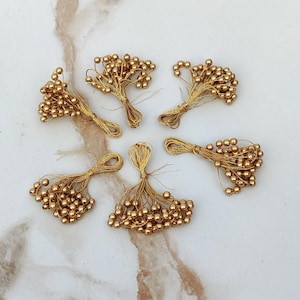 May include: Five bundles of gold-coloured artificial flower stamens, each consisting of numerous small, round beads attached to thin, metallic gold-toned wire stems. The clusters are arranged in a circular pattern, showcasing the shiny, reflective surface of the beads and the delicate, flexible wire construction of these craft components.