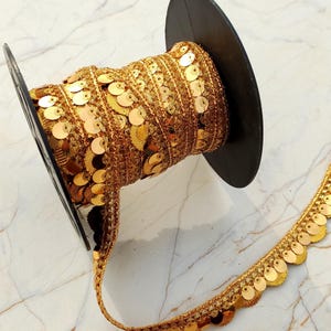 May include: A spool of gold metallic trim featuring a decorative scalloped edge adorned with small, shiny circular sequins. The trim is wrapped around a black plastic reel, showcasing a dense, textured weave with a reflective finish throughout.