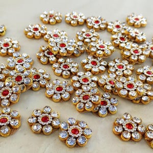 Puede incluir: Una colección de pequeños adornos en forma de flor con una piedra central roja rodeada por seis diamantes de imitación blancos transparentes. Cada pieza está montada en un marco de metal dorado, creando un diseño floral uniforme en todos.