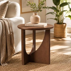 May include: A round wooden side table with a dark brown finish and a curved, open-frame base. A beige ceramic vase containing green leafy branches and a book titled KINFOLK are placed on the flat circular tabletop surface.