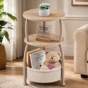May include: A three-tier round side table with a light wood grain finish and curved metal legs. The top shelf holds a green succulent in a white textured pot. The middle shelf features a clear acrylic sign with the text Tomorrow es never. The bottom shelf includes a fabric basket containing a tan teddy bear holding a pink heart and two books.
