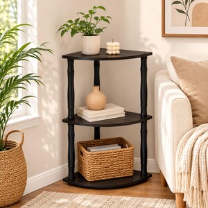 May include: A three-tier black corner shelf unit with a triangular shape. The unit features three dark wood-grain shelves supported by four black cylindrical pillars. The shelves hold a small potted plant, a candle, a vase, books, and a basket.