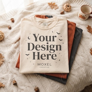 Puede incluir: Una pila de tres camisetas dobladas en color crema, naranja quemado y gris carbón. La camiseta superior de color crema presenta un texto gris oscuro que dice Your Design Here y MOXEL CREATIVE, rodeado de pequeñas ilustraciones gráficas de murciélagos, estrellas y lunas.