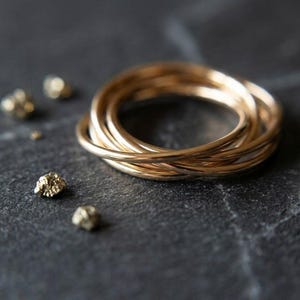 May include: A gold-colored ring composed of five thin, interlocking bands that create a layered, circular structure. The ring is accompanied by several small, irregular, metallic gold-colored nuggets scattered nearby on a dark, textured surface.