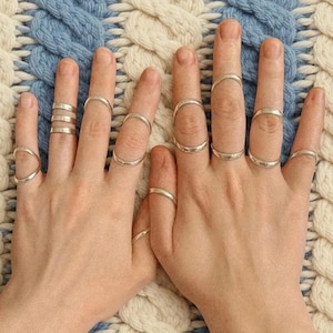 Può includere: Una collezione di dodici anelli color argento indossati su entrambe le mani. Gli anelli presentano fasce semplici, lisce e sottili, con alcuni posizionati alla base delle dita e altri indossati come anelli midi tra le nocche di varie dita.