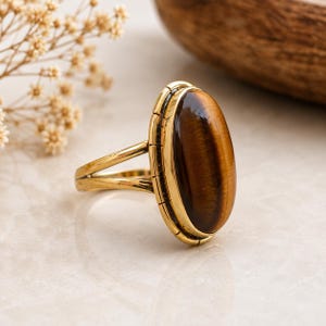 Puede incluir: Un anillo de tono dorado con una gran piedra preciosa de ojo de tigre de forma ovalada engastada en un bisel liso. La banda se divide en dos hilos finos y paralelos que se conectan a los lados del engaste, con un detalle de borde dentado.