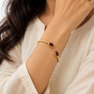 May include: A gold-toned open cuff bracelet featuring a hammered metal texture and two teardrop-shaped dark red garnet gemstones at each end. The bracelet is designed as a flexible open band that rests on the wrist with the stones facing forward.