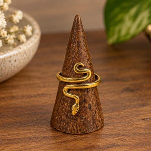 Könnte beinhalten: Ein goldfarbener Metallring in Form einer gewundenen Schlange mit detailliertem Kopf und einem schlanken Körper, der sich um das Band legt. Der Ring wird auf einem dunkelbraunen, kegelförmigen Holzständer präsentiert, der das Metallfinish betont.