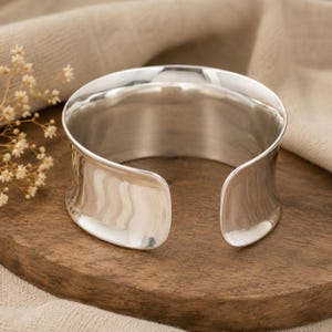 Pode incluir: Uma pulseira larga de prata polida com uma superfície lisa e reflexiva e um design curvo aberto. A pulseira apresenta uma largura consistente ao longo de toda a sua forma circular, exibindo um acabamento metálico limpo e bordas arredondadas.