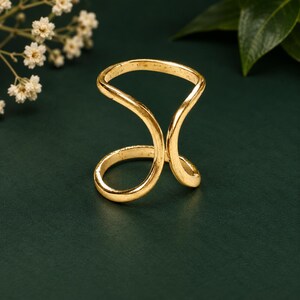 Op de afbeelding: Een goudkleurige ring met een open bypass-ontwerp, bestaande uit twee gebogen, parallelle banden die in het midden samenkomen. Het metaal heeft een gladde, gepolijste afwerking en vormt een vloeiende, doorlopende vorm rond de vinger.