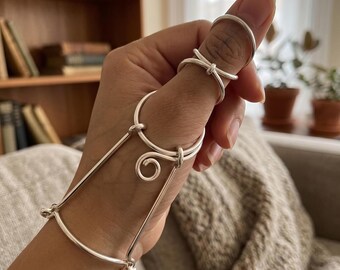 Pode incluir: Uma tala de dedo em metal prateado com um anel circular em torno do polegar e uma estrutura conectada que se estende até uma pulseira. O design inclui um elemento decorativo em espiral e componentes de fio polido e liso.