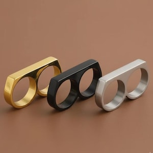 May include: Three double-finger rings in gold, black, and silver tones. Each ring features a smooth, polished metal finish with two circular openings for fingers connected by a single, solid, rectangular bar across the top surface.
