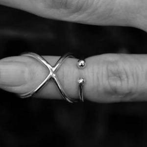 May include: A silver-toned ring featuring a crossover X-shaped design that wraps around the finger, ending in two small, smooth spherical beads at the open gap. The metal has a polished, reflective finish throughout the entire structure.