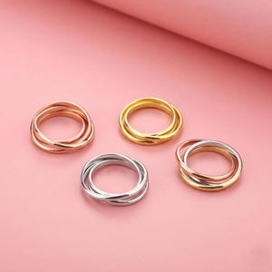 Può includere: Quattro anelli intrecciati, ciascuno composto da tre sottili fasce con diverse finiture metalliche. Il set include combinazioni di tonalità oro rosa, oro giallo e argento, disposte in una formazione circolare con superfici lisce e lucide.