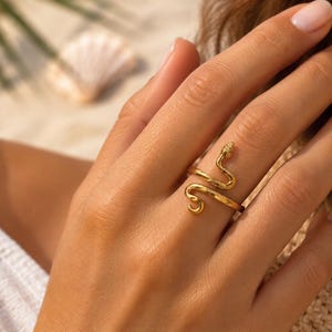 Op de afbeelding: Een goudkleurige ring in de vorm van een kronkelende slang. De metalen band wikkelt zich om de vinger, met een gedetailleerde kop aan het ene uiteinde en een gekrulde staart aan het andere. Het oppervlak van de ring heeft een gladde, gepolijste afwerking.