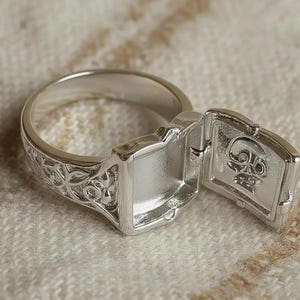 Può includere: Un anello in metallo color argento caratterizzato da un intricato motivo in filigrana lungo la fascia e da uno scomparto incernierato a forma di scatola sulla parte superiore. Il coperchio è aperto, rivelando un piccolo teschio in rilievo.