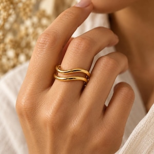 Puede incluir: Un anillo de tono dorado con un diseño de doble banda y una forma ondulada suave. Las dos bandas paralelas están conectadas, creando una silueta continua y fluida que envuelve el dedo con un acabado pulido y reflectante.