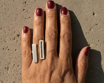 May include: A silver ring featuring two distinct rectangular bars positioned vertically across the finger. One bar is solid and smooth, while the other bar contains a thin, recessed vertical channel running through its center, creating a geometric design.